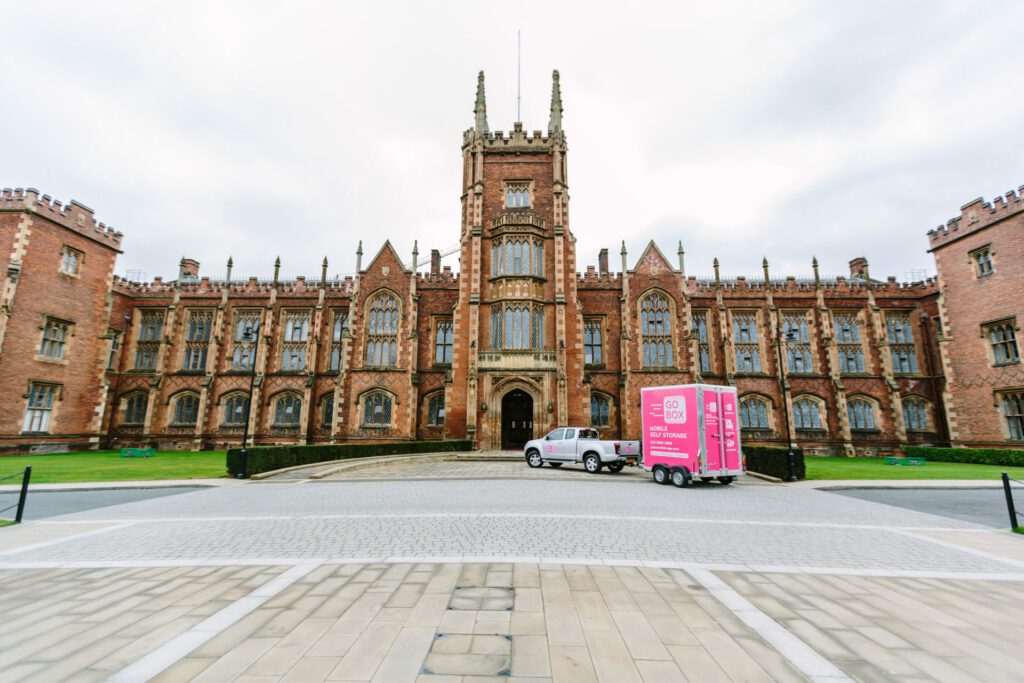 A pick-up truck towing a mobile storage unit in front of a historic gothic-style building under a cloudy sky.