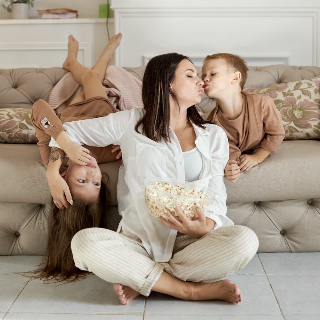 A mother sits on the floor holding a bowl of popcorn while receiving kisses from her son and daughter during a relaxed family moment at home.