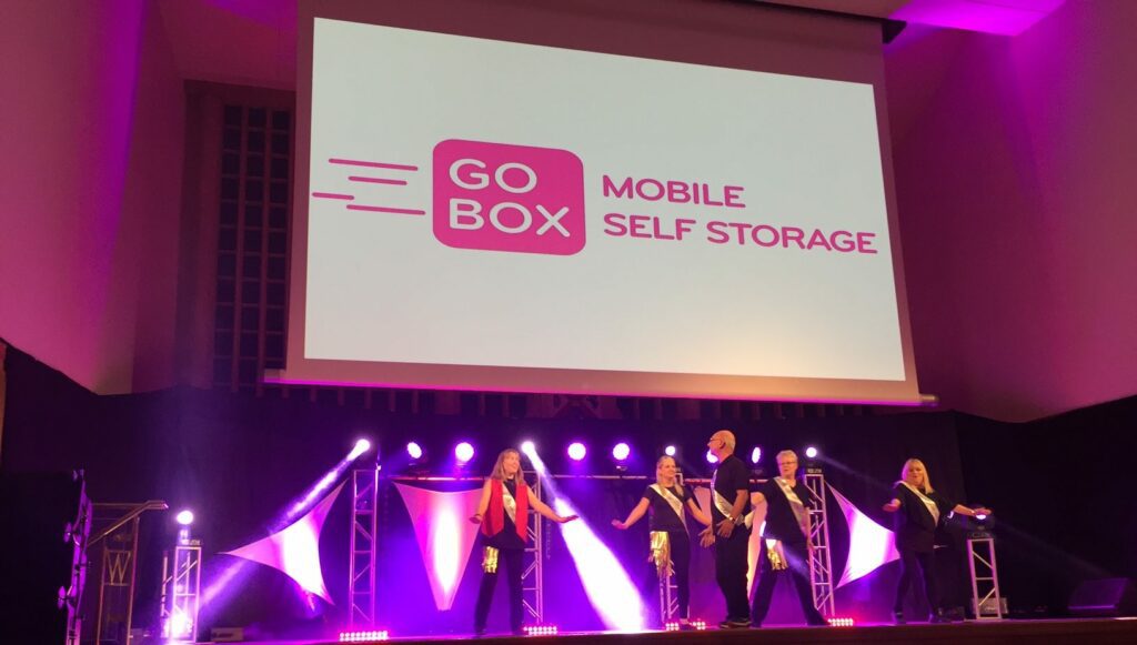 A group of individuals performing on stage at an event featuring a backdrop with the 