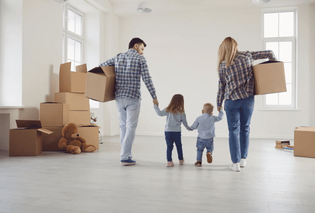 A family carrying boxes into a new home with unpacked boxes and a teddy bear in the background.