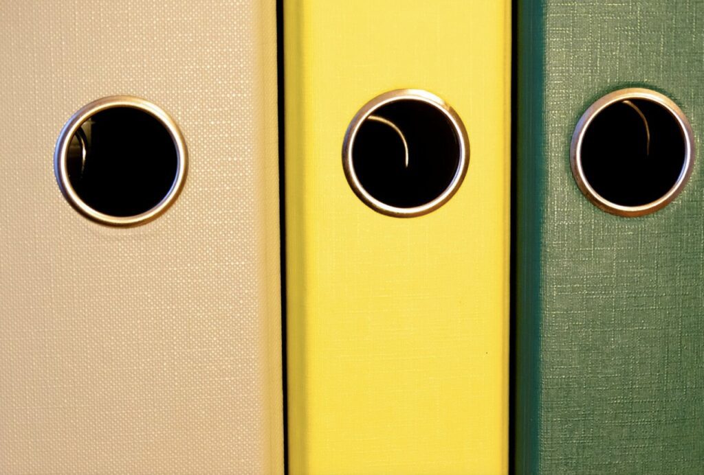 Three binders with black rings on their spines, aligned vertically, in pastel shades of beige, yellow, and green.