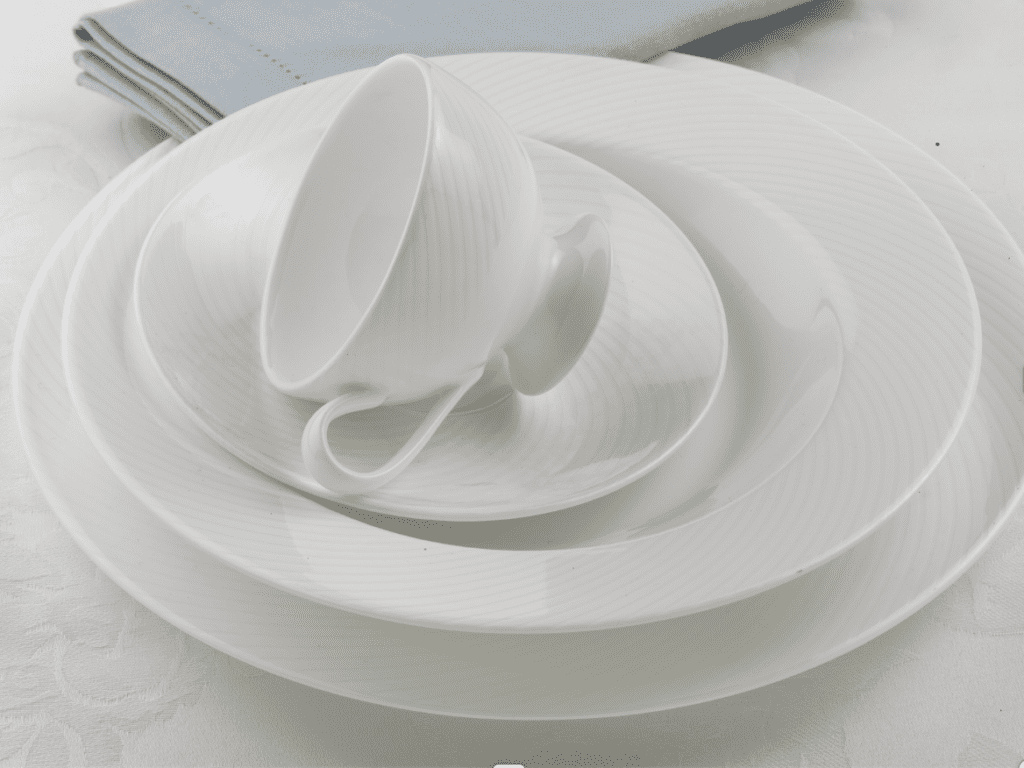A stack of clean white dinnerware with a toppled cup on top.