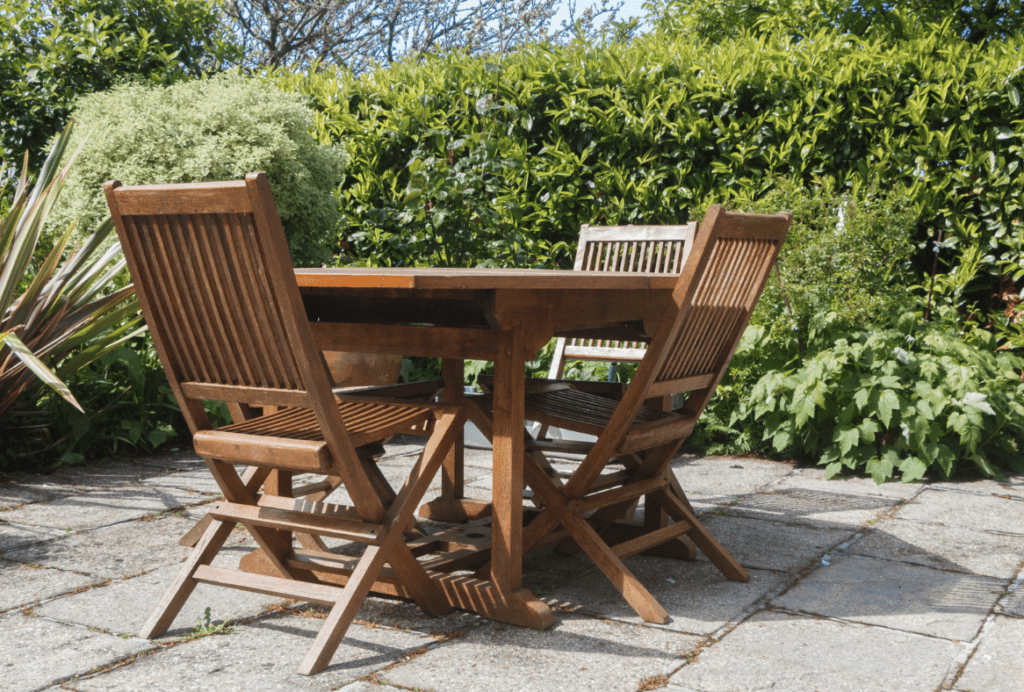 Wooden patio furniture consisting of a table and chairs set on an outdoor paved area with surrounding greenery.