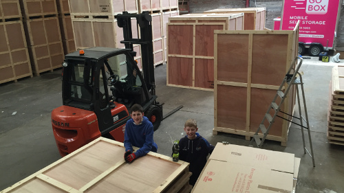 Two children posing in a warehouse with wooden crates and a forklift in the background.