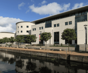 Modern office building façade reflecting on a calm waterfront under partly cloudy skies.