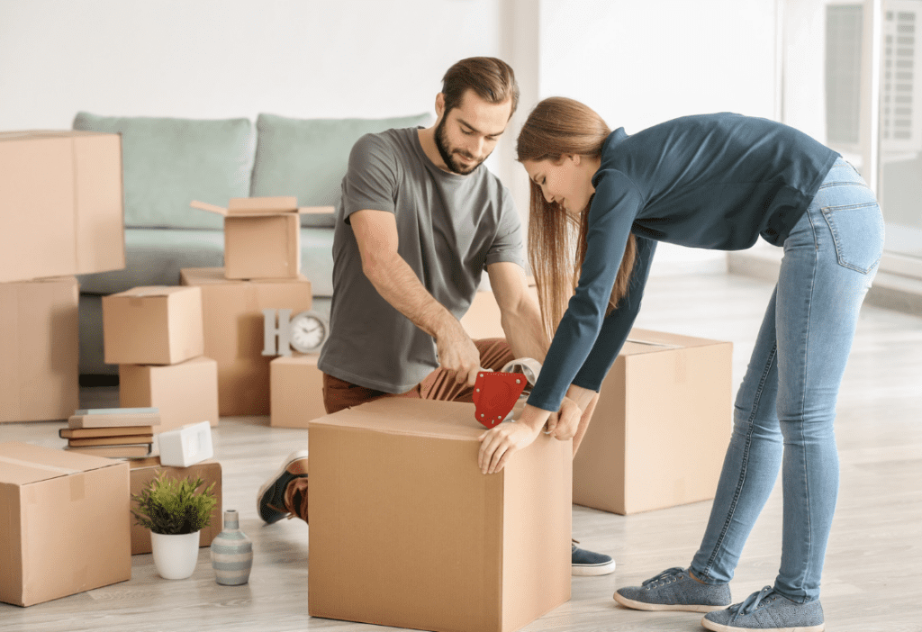 Two people packing items into boxes in a room with unpacked boxes and furniture.