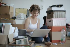 Woman smiling while using a laptop amidst moving boxes in a cluttered room.