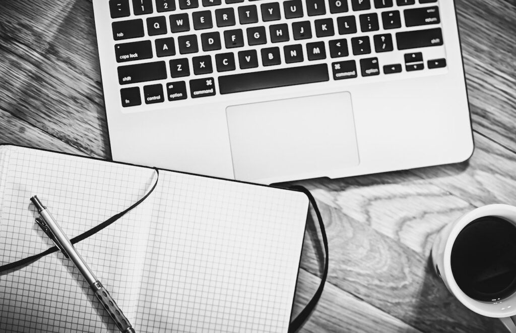 Black and white photo of a laptop, notepad with a pen, and a cup of coffee on a wooden desk in a working from home environment.