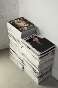 A stack of assorted magazines, featuring essential ways to successfully sell your home, placed on a white block against a white brick wall.