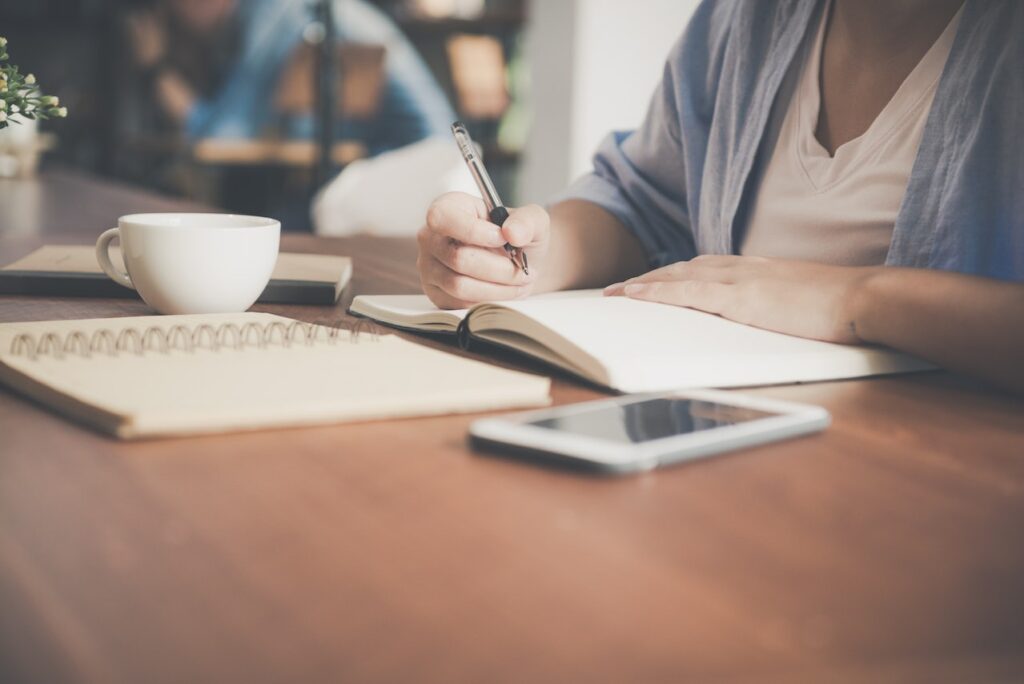 Person working from home, writing in a notebook at a table with a coffee cup and smartphone nearby.