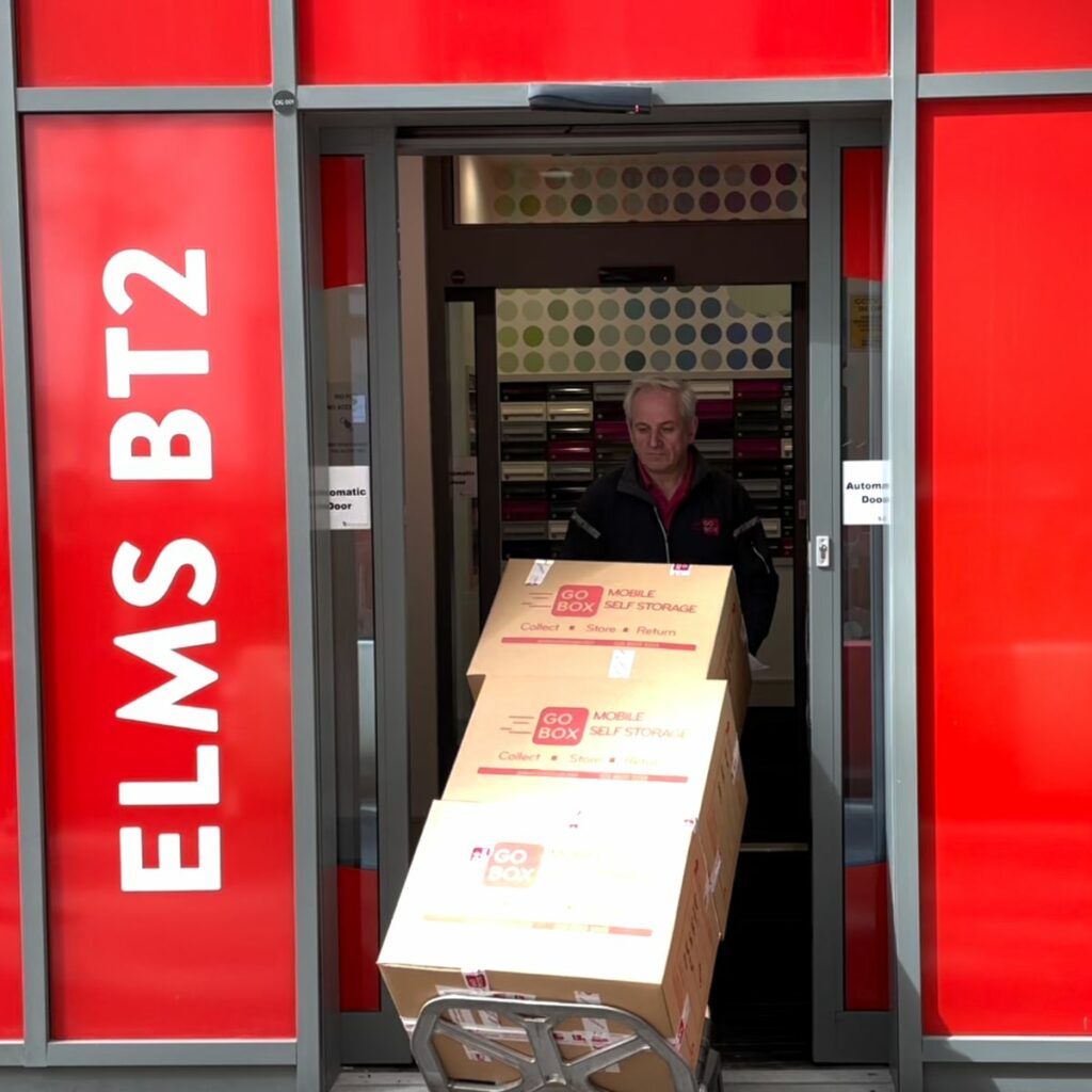 A person in a red jacket using a trolley to transport large boxes out of a building with red signage.